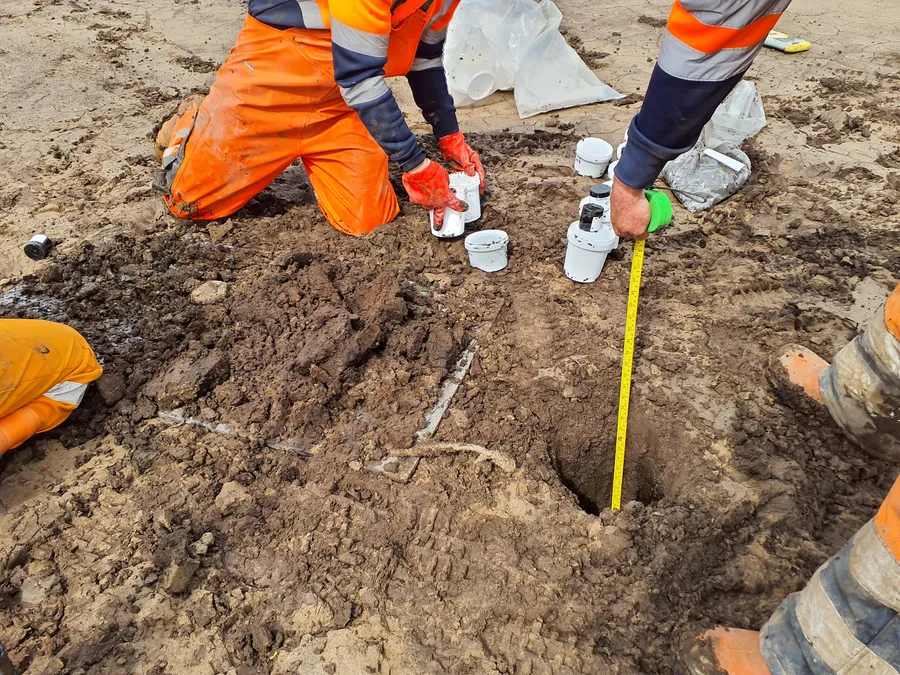 Site workers in orange hi-vis kneeling by a small pit, measuring it with a tape and taking samples.