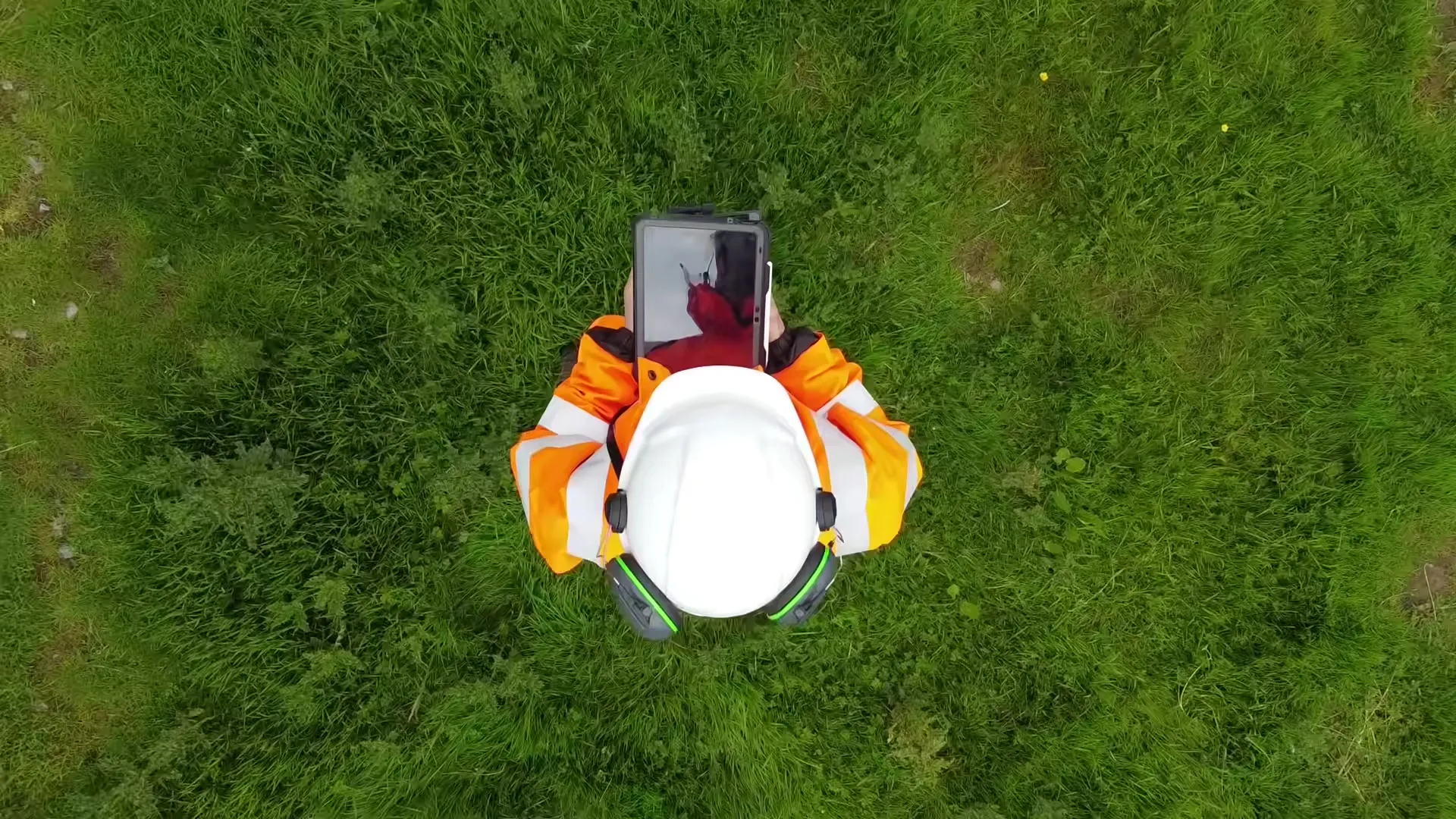 Drone view straight down on a surveyor in hi-vis holding a tablet in a grass field.